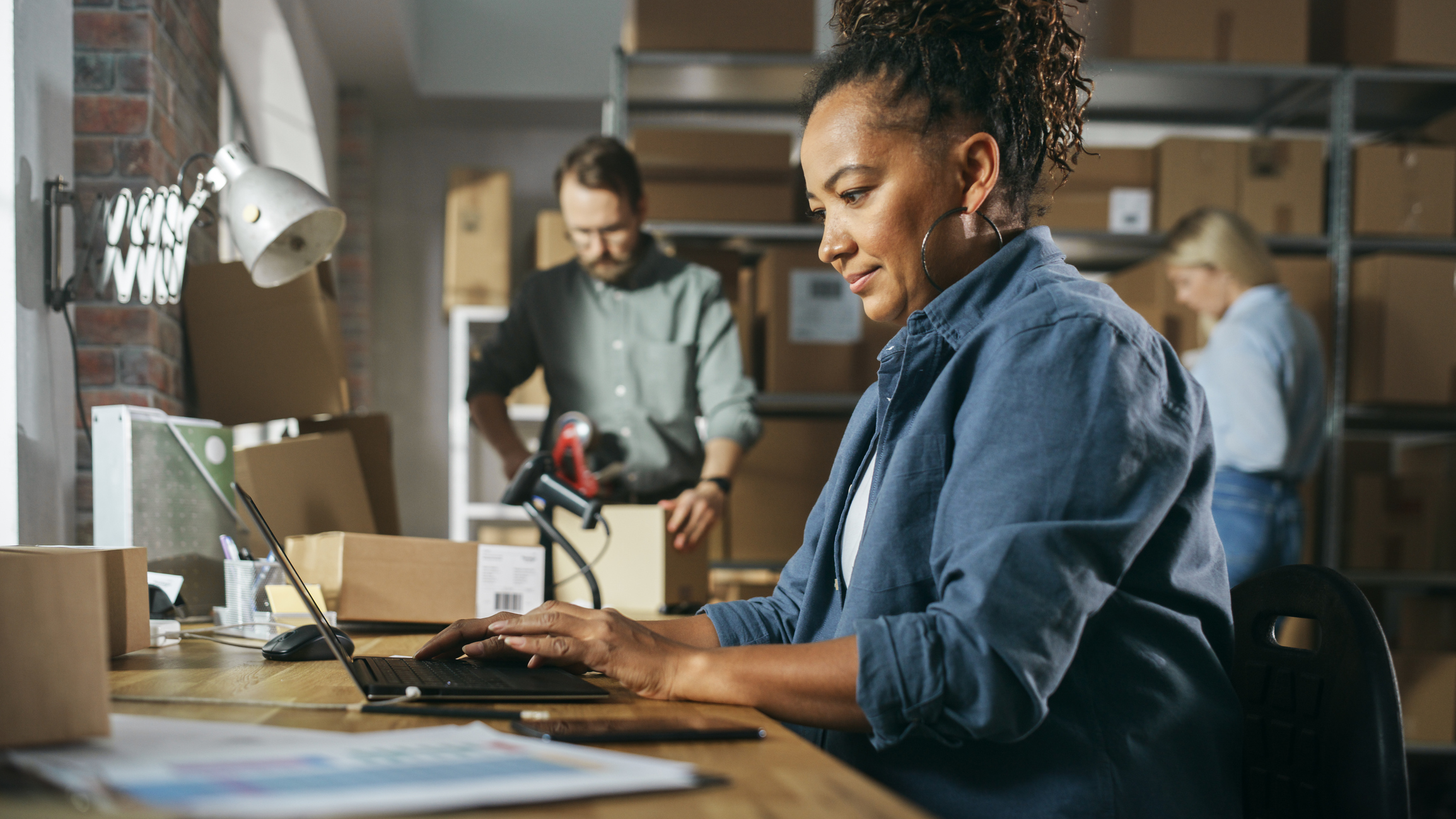 Woman using laptop in a back storage room with two colleagues behind her.